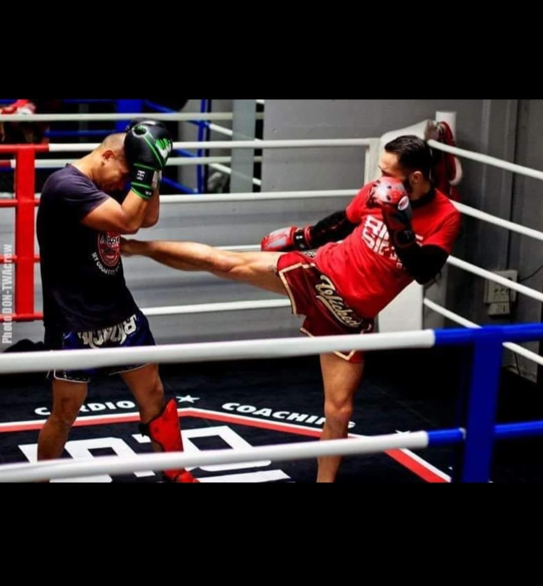 Coach de Muay Thaï torse nu posant dans une cage d’entraînement, équipé de paos rouges et bleus prêts pour une séance.